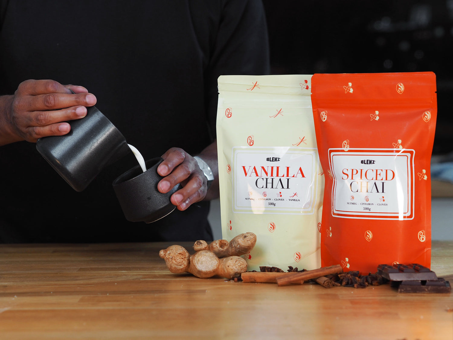 Bags of Vanilla and Spice Chai Powder on cafe bench with barista pouring steamed milk